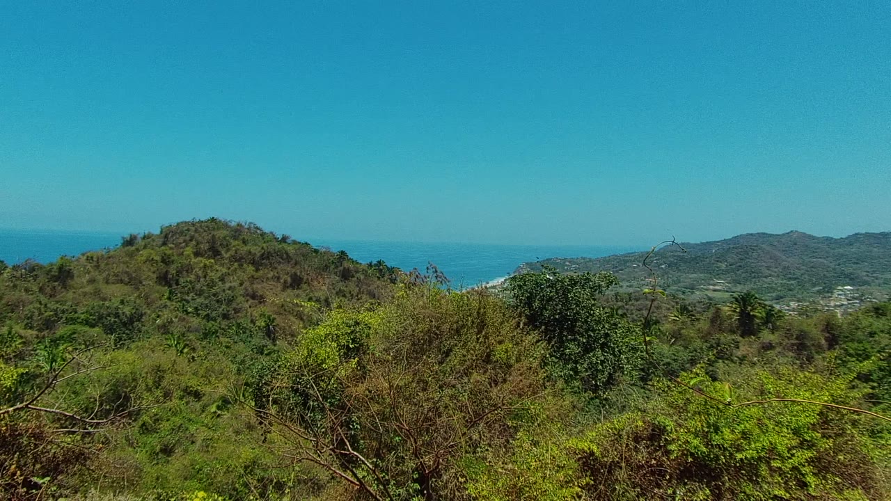 Early frame of a San Pancho, Nayarit hillside covered in green vegetation, with blue ocean and a clear horizon behind it and no visible people.