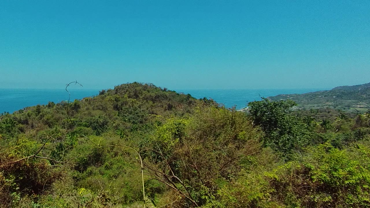 San Pancho coastal greenery spreads across the foreground while the hill rises in the center and strips of ocean appear near both sides, with no visible people.