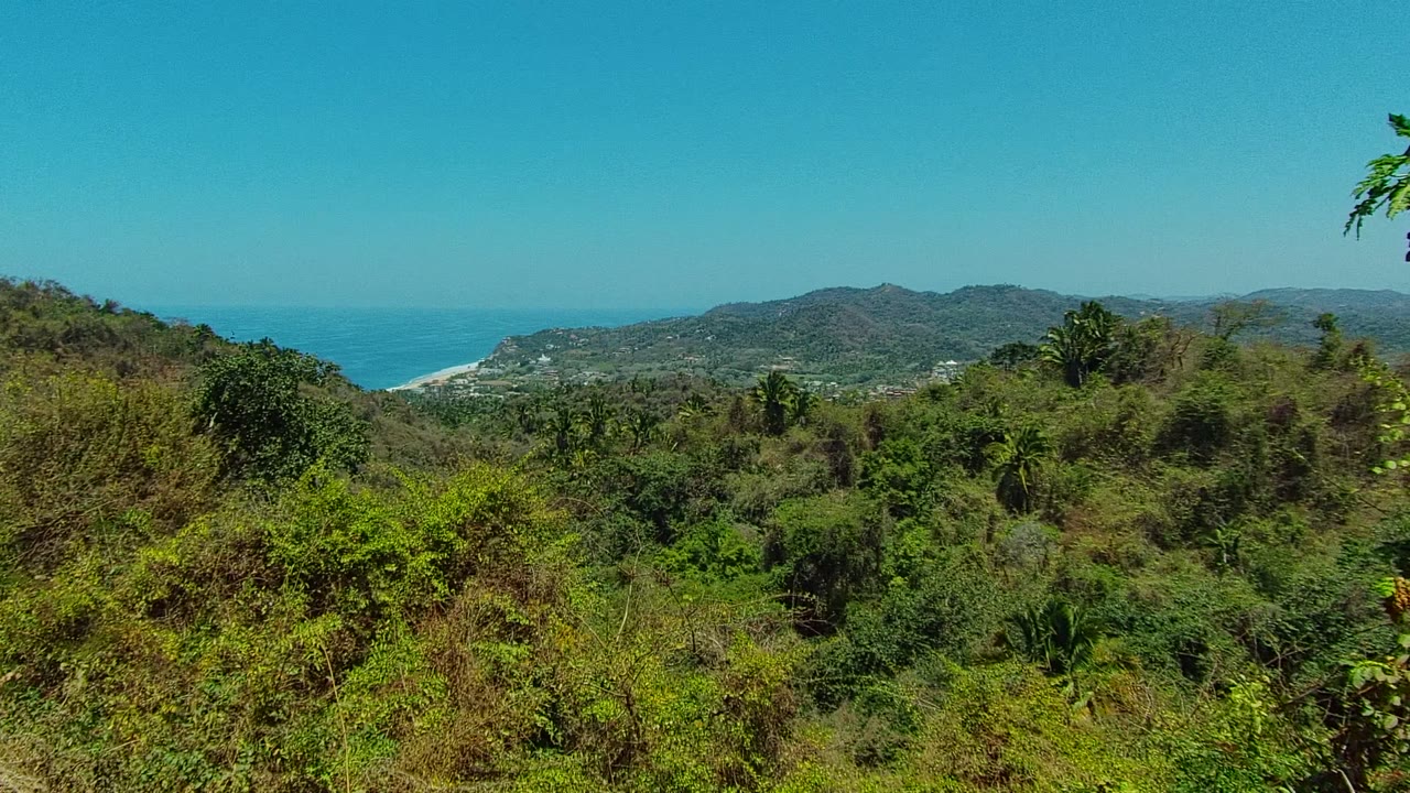 Wide elevated view over green and dry hillside vegetation near San Pancho, Nayarit, with distant mountains, a strip of beach, blue ocean, clear sky, and no visible people.