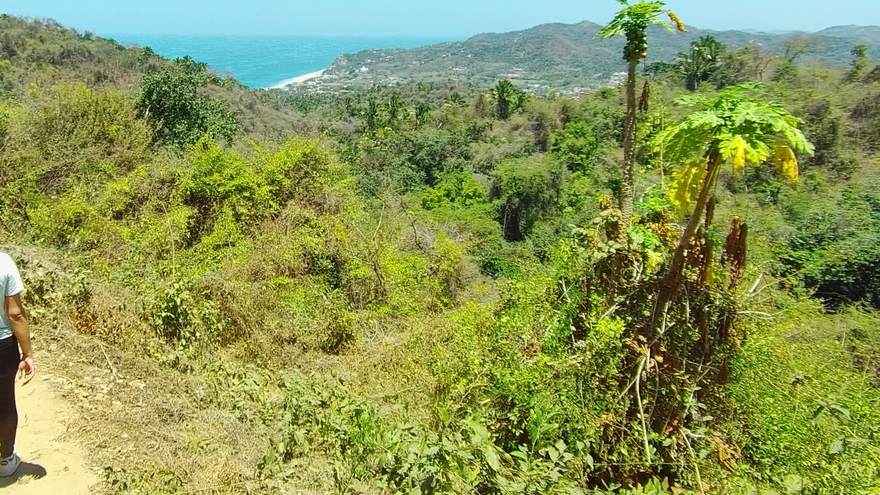 A hiker is partly visible at the left edge of a San Pancho, Nayarit dirt trail, with dense hillside vegetation, rounded hills, blue ocean, and clear sky in the distance.