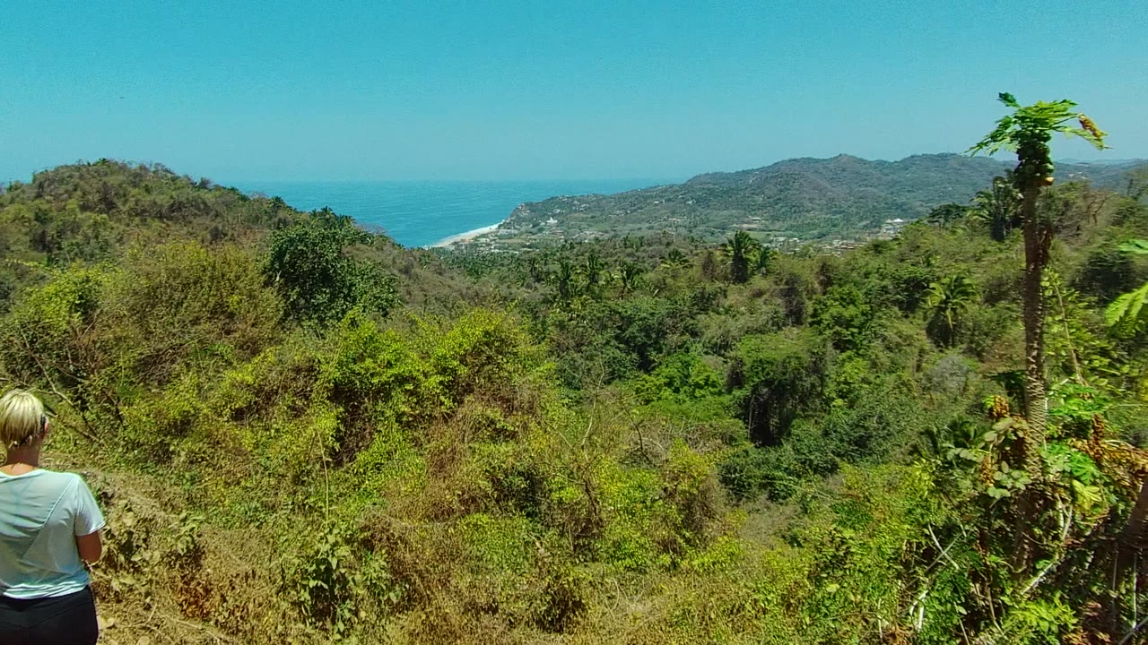 The hiker appears lower in the San Pancho trail frame as vegetation fills the foreground, with hills, a pale surf line, blue ocean, and clear sky in the background.