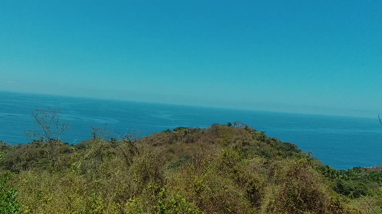 San Pancho, Nayarit hillside brush and a rounded green-brown hill overlook the blue ocean under a clear sky, with no visible people.