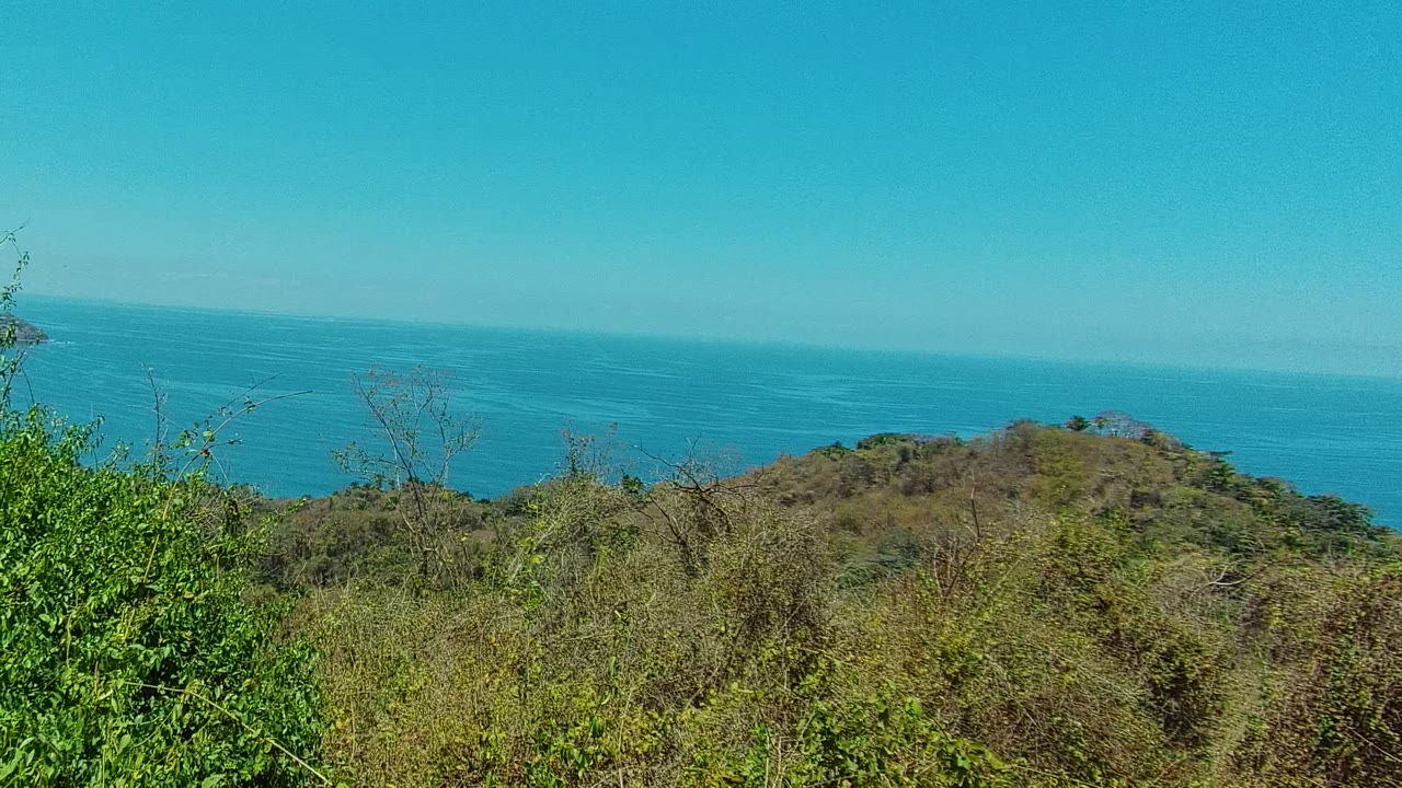 Opening frame of a San Pancho, Nayarit hilltop view with green foreground brush, a rounded coastal hill, blue ocean on the horizon, and no visible people.