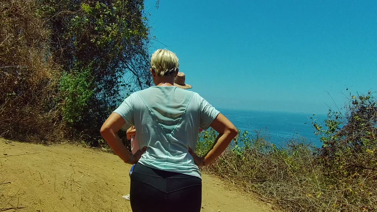 In San Pancho, Mexico, an adult in a light shirt stands foreground on a dusty coastal trail while brush frames a blue ocean horizon.