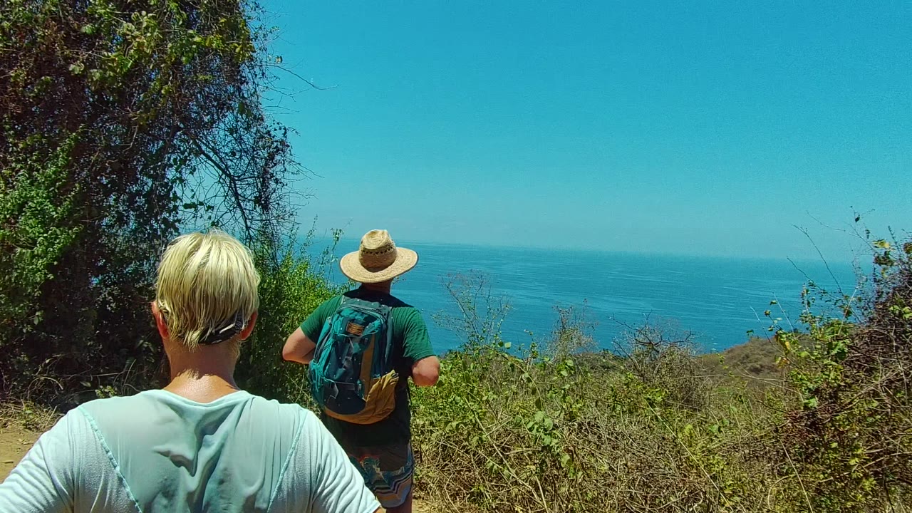 In San Pancho, Mexico, the foreground adult is seen from behind while a hat-wearing hiker with a backpack faces the ocean over low brush.