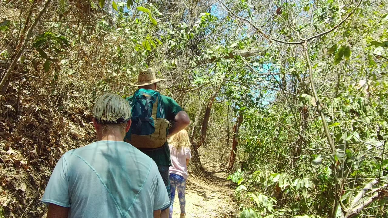 Two adult hikers and a child are visible from behind on the San Pancho trail, with bright sun on the path and dense green foliage on both sides.