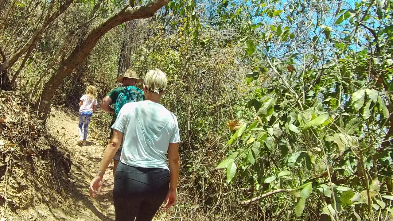 Three hikers walk away from camera on a narrow dirt trail in San Pancho, Nayarit, Mexico, with a child ahead and two adults closer to the foreground.