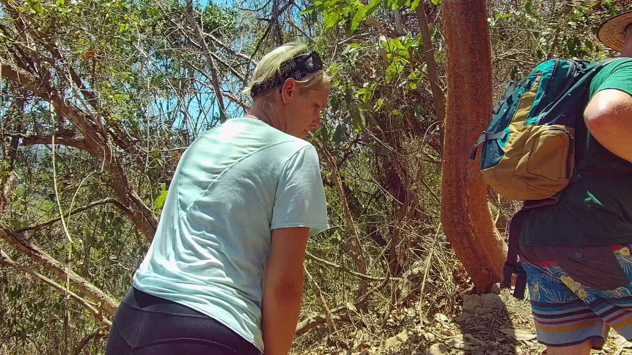 Two hikers move up a narrow dry forest trail in San Pancho, Mexico, with a pale-shirted hiker in the left foreground and a backpacked hiker ahead.