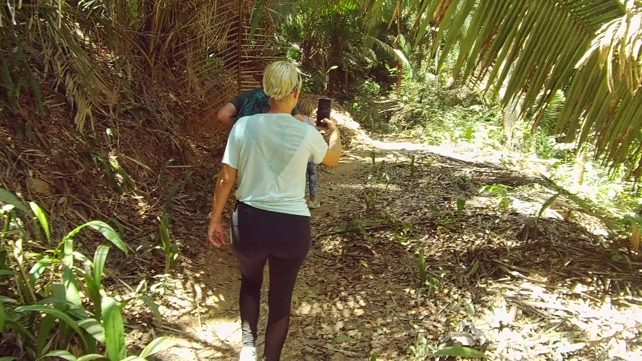 An adult on the right side of the San Pancho trail holds up a phone while walking, with a second person partly visible ahead and bright foliage lining the path.