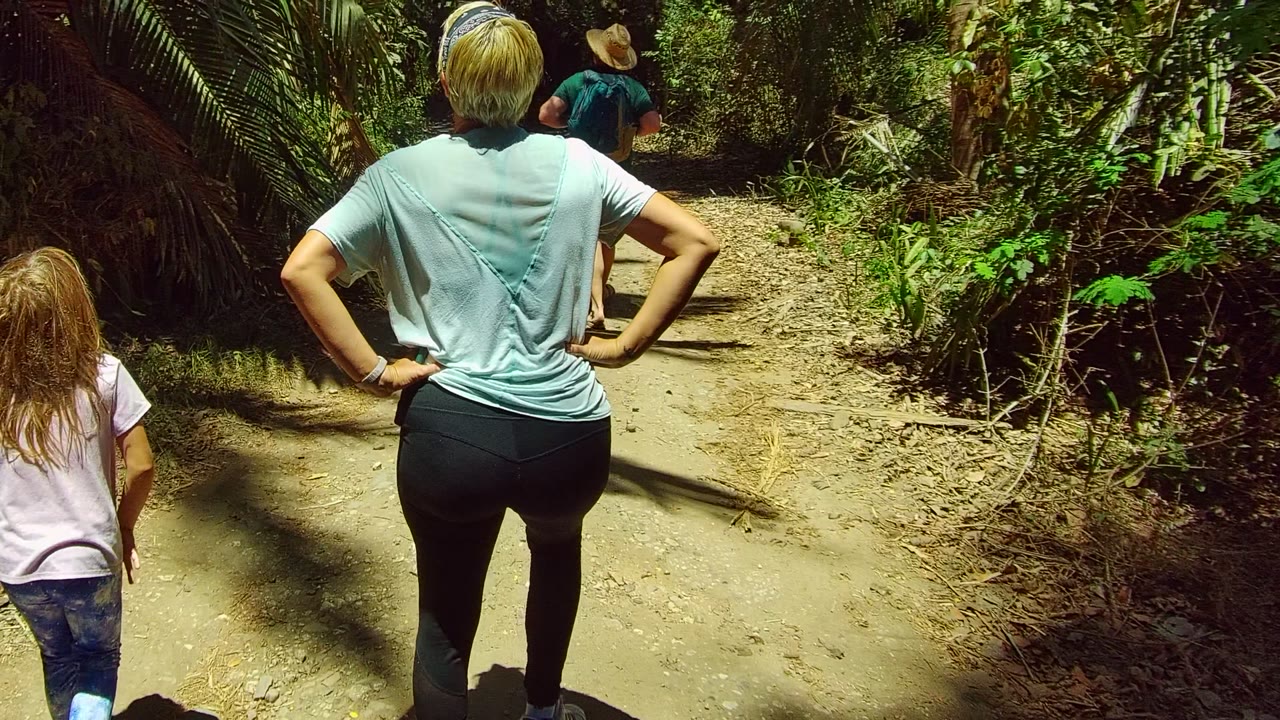The adult and child continue down the San Pancho forest trail while a hiker in a hat walks ahead between dense green plants.