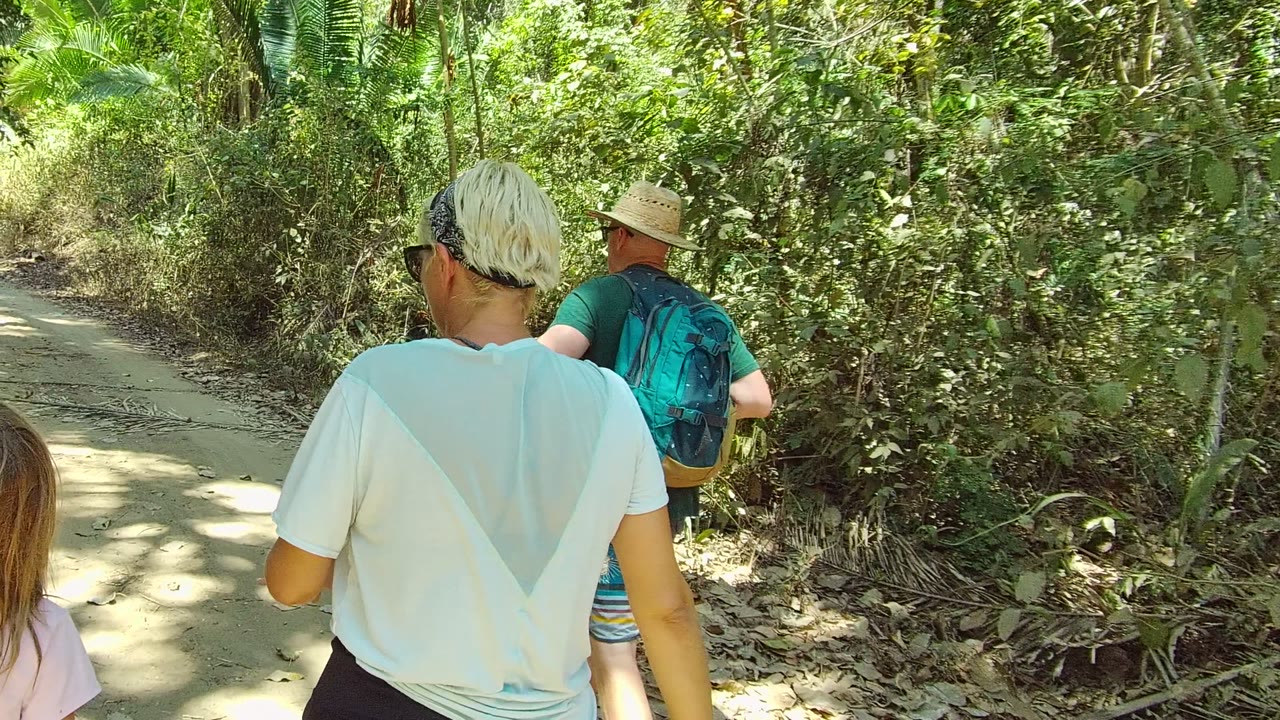 People walk away from the camera on a shaded dirt trail in San Pancho, Mexico, with a blue backpack, straw hat, and dense green vegetation visible.