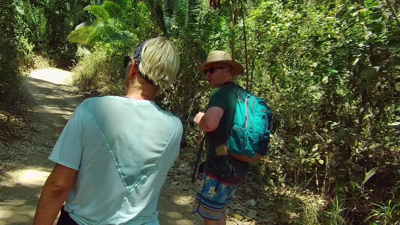 A man with a straw hat and blue backpack walks ahead on a narrow San Pancho forest trail while another person follows near the camera.