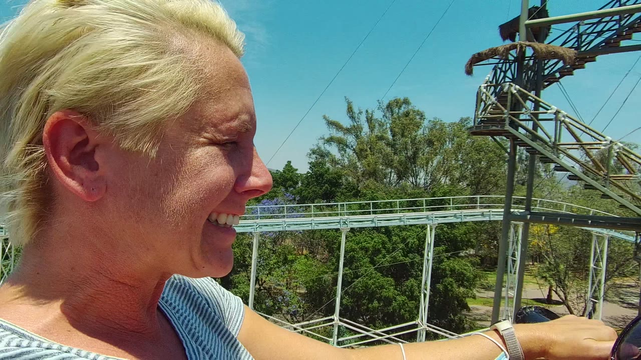 The blonde rider smiles in profile beside the safety bar in Guadalajara, Mexico, with a roller coaster support tower, pale blue track, trees, and sky in the background.