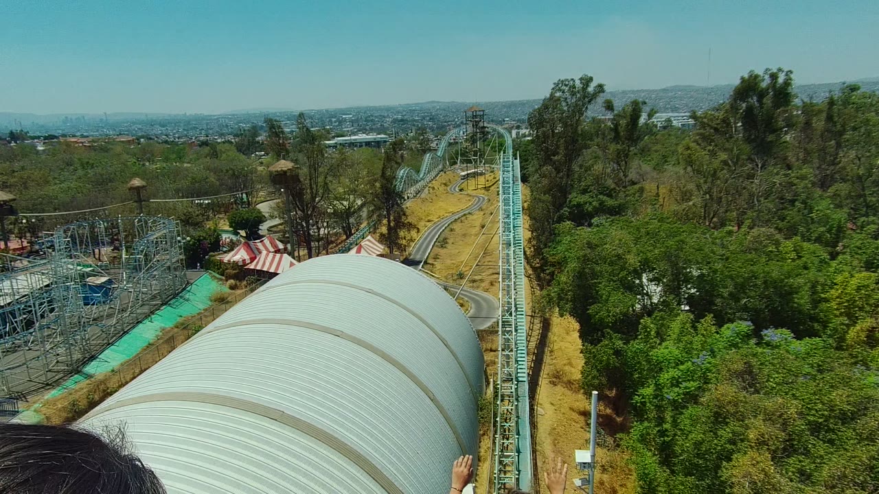 Wide forward view from a roller coaster in Guadalajara, Mexico, with turquoise track, a large silver ride structure, trees, distant city horizon, and partial passengers at the bottom edge.