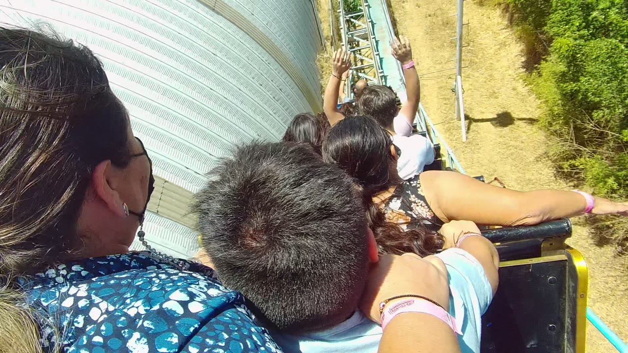 Passengers ride a roller coaster beside a ribbed silver structure with hands raised ahead.
