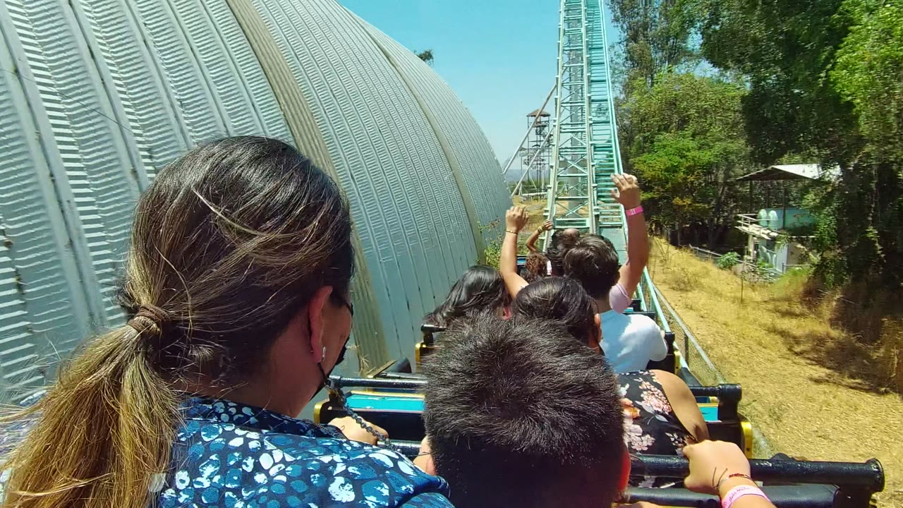 Rear passenger view on a Guadalajara, Mexico roller coaster, with a woman and child in the foreground, riders ahead, turquoise track rising in the distance, dry grass, green trees, and clear sky.