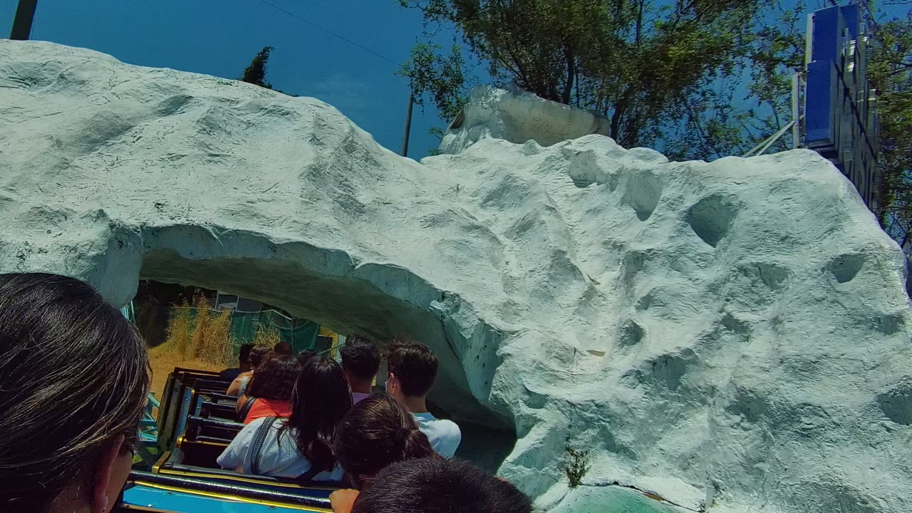 Passengers on a roller coaster in Guadalajara, Mexico pass beneath a pale artificial rock arch, with heads and seat backs filling the lower foreground.
