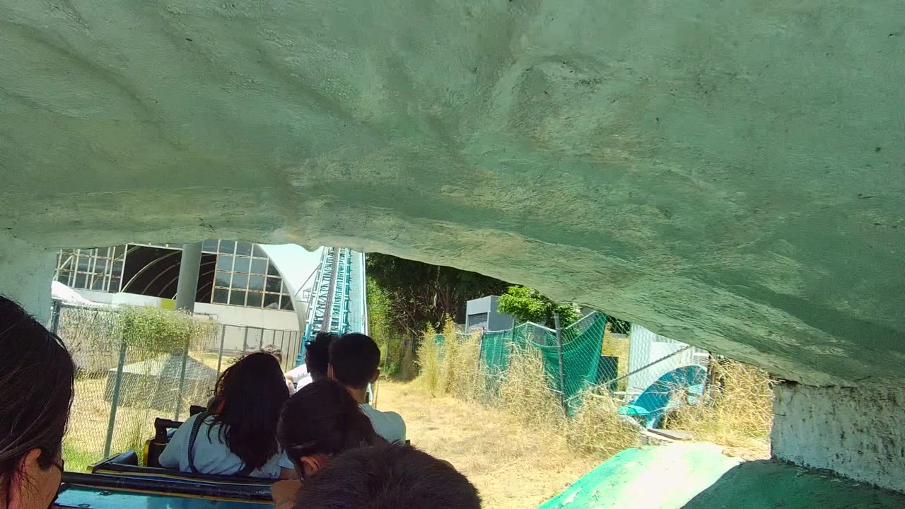 Roller coaster riders emerge from under a rock overhang toward park structures.