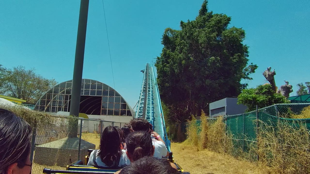 Passengers continue forward on the roller coaster in Guadalajara, Mexico, with a blue track rail, chain-link fence, tall trees, and a dome structure visible ahead.