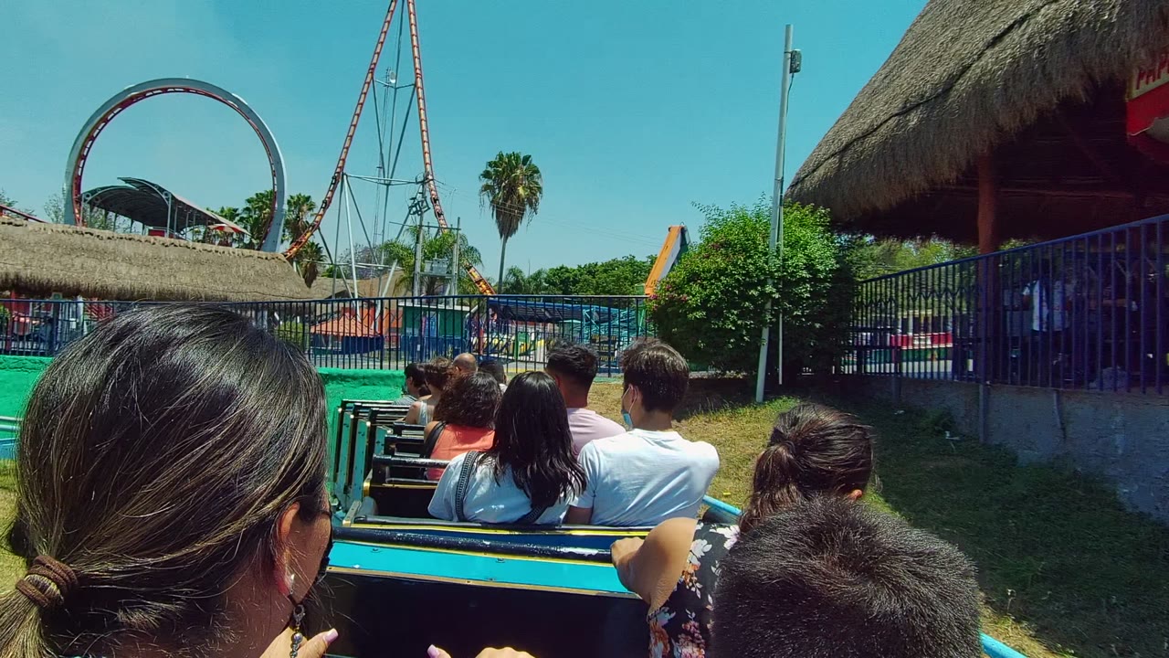 Forward ride view in Guadalajara, Mexico with passenger heads in the foreground, a blue fence, thatched roofing, green netting, and a roller coaster loop beyond.