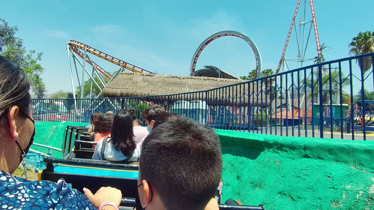 A closer foreground view shows passenger heads and hands inside the ride in Guadalajara, Mexico, beside a green wall, blue fence, thatched roof, and looping coaster track.