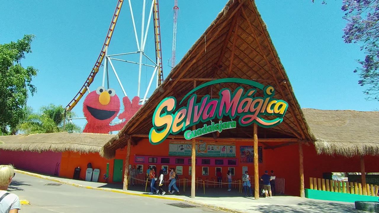 Wide view of the Selva Mágica Guadalajara entrance in Guadalajara, Mexico, with the thatched roof sign centered, visitors near the ticket windows, and an Elmo figure partly visible behind the wall.
