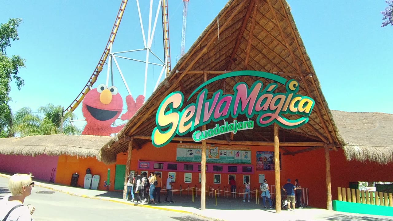 The Selva Mágica Guadalajara facade remains centered in Guadalajara, Mexico, with the sign spanning the thatched roof, a small group at the entrance, the Elmo figure behind the wall, and coaster track overhead.