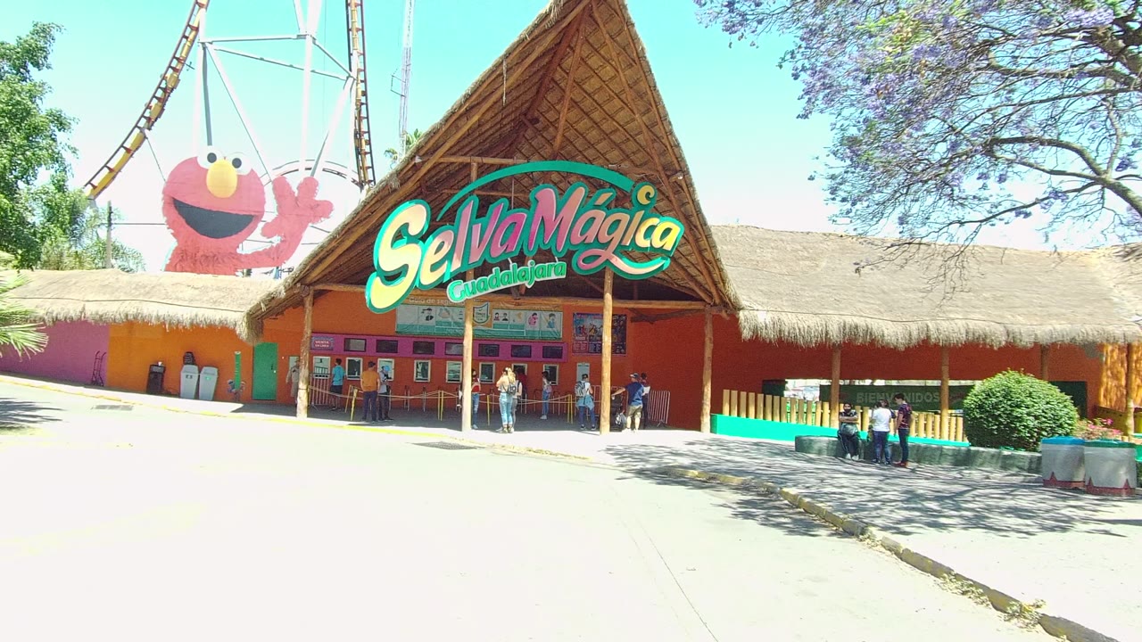 Wide frame of the Selva Magica theme park entrance in Guadalajara, Mexico, with the thatched roof sign centered, the Elmo display to the left, and visitors near the doors.