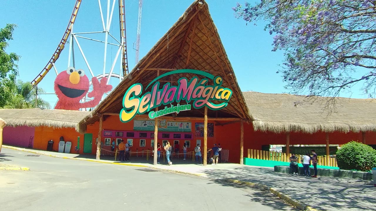 The Selva Magica entrance remains in a wide daytime view, with the Elmo display left of the roof peak and several visitors gathered near the shaded doorway area.