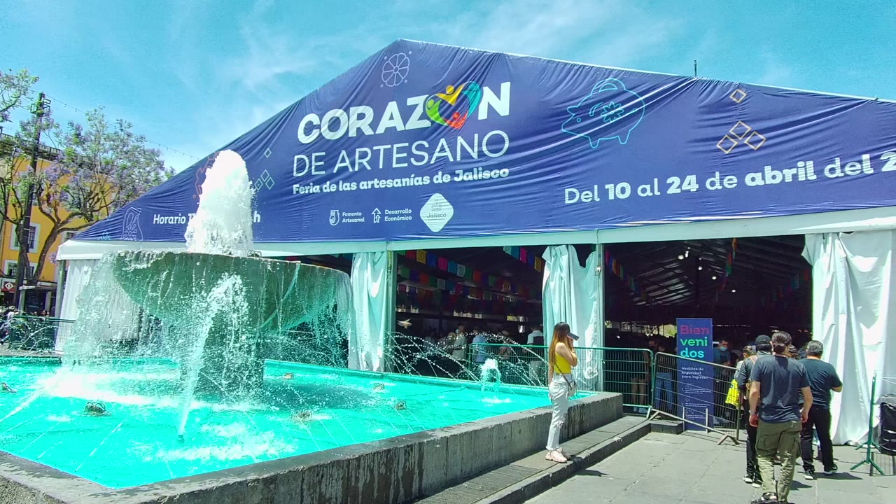 A turquoise fountain sprays in front of the blue Corazon de Artesano artisan fair tent in Guadalajara, Mexico, with several people standing and walking near the entrance.