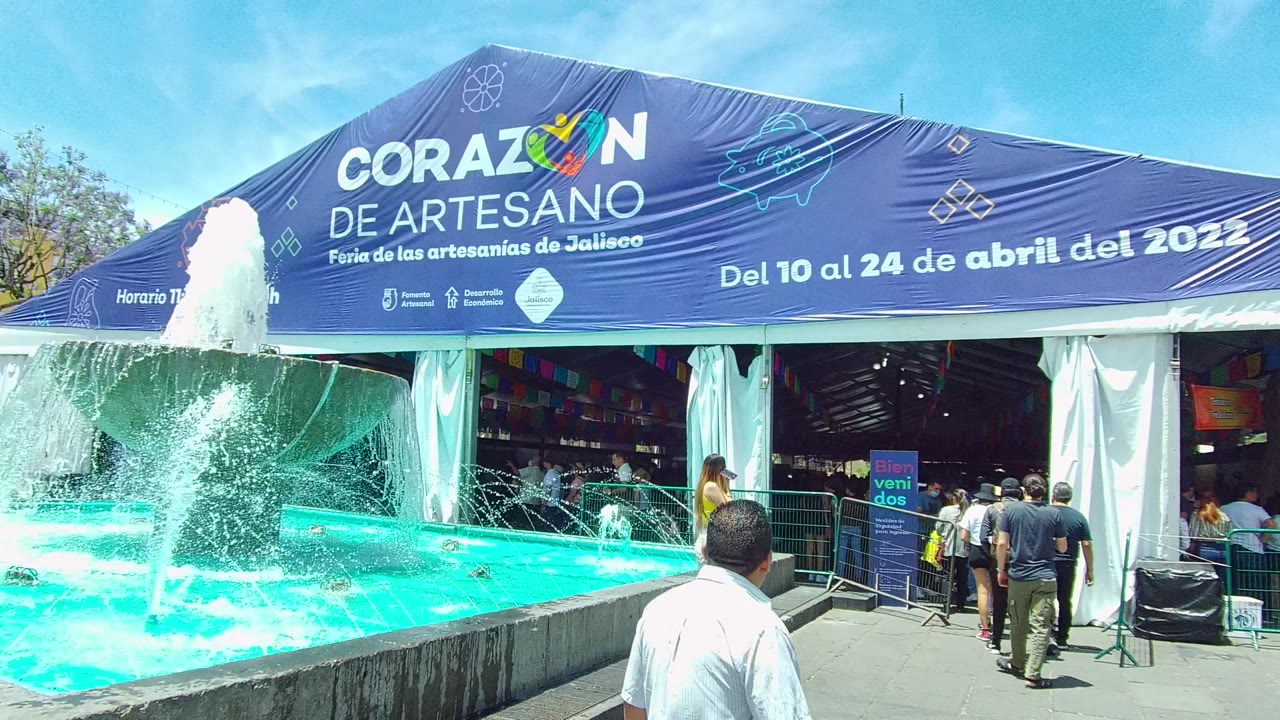 The fountain spray shifts across the turquoise pool as a man stands near the lower frame edge and people gather at the blue Corazon de Artesano tent in Guadalajara, Mexico.