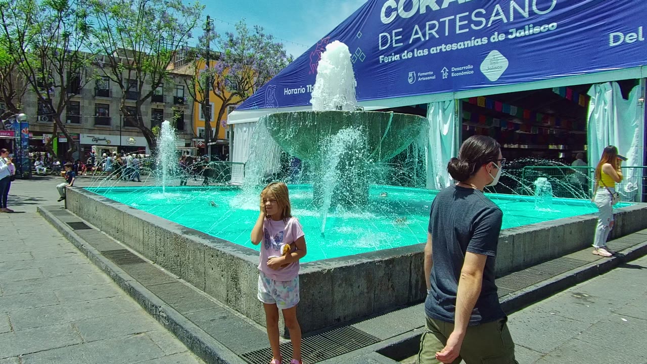 Wide view in Guadalajara, Mexico of a blue-water fountain in front of a CORAZON DE ARTESANO tent, with a child near the foreground and adults around the basin.