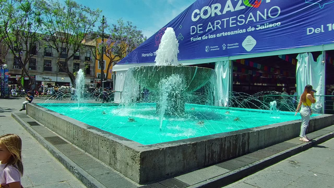 A wide Guadalajara market view holds the turquoise fountain across the foreground, with a woman standing near the tent and a child partly visible at the bottom right.