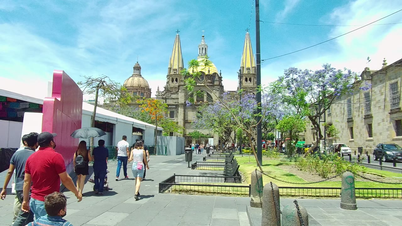 Pedestrians walk along a Guadalajara, Mexico city square, with a pink structure at left, benches and trees ahead, and cathedral spires in the background.