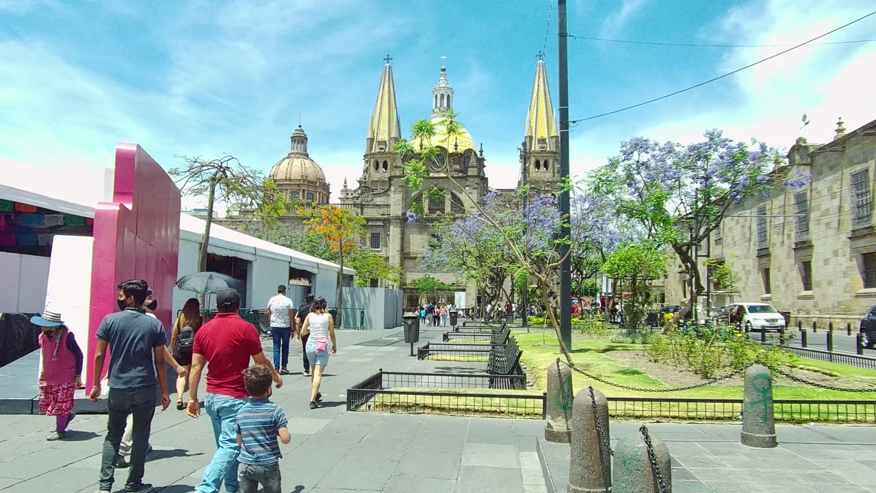 People walk past a pink structure beside a plaza and cathedral.