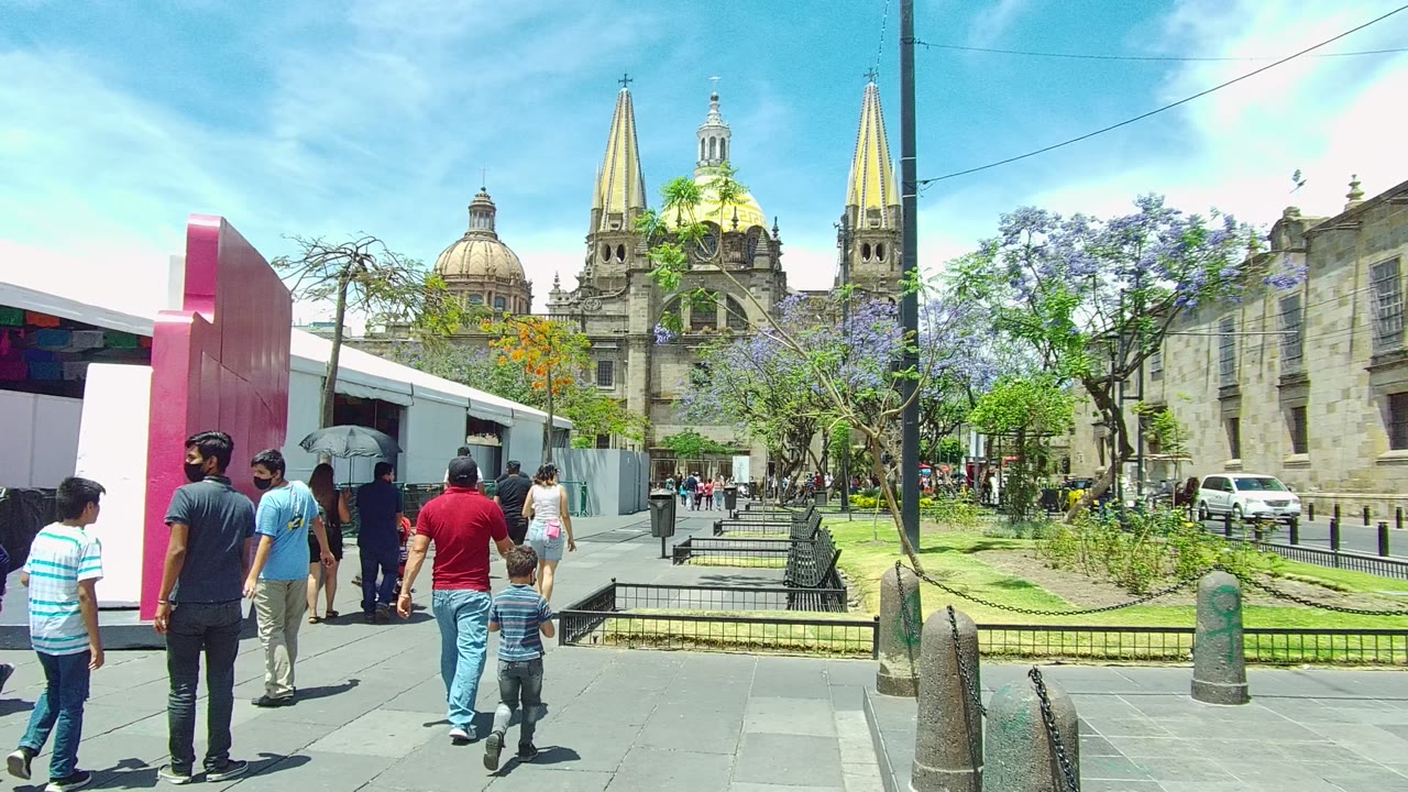 Pedestrians including a child walk across the foreground of the Guadalajara city square while the cathedral dome, twin spires, benches, and flowering trees remain in view.