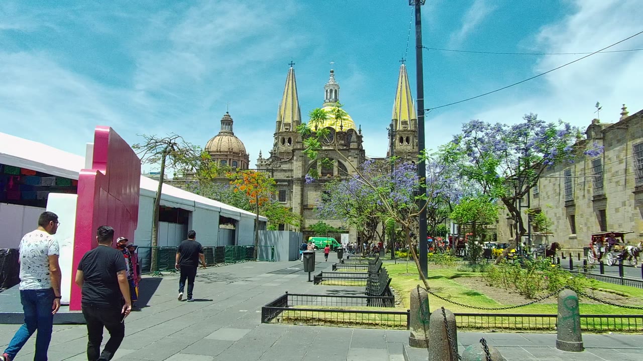 Pedestrians walk through a paved plaza in Guadalajara, Mexico, with a pink structure at left, benches and trees in the middle, and a cathedral with golden spires in the background.