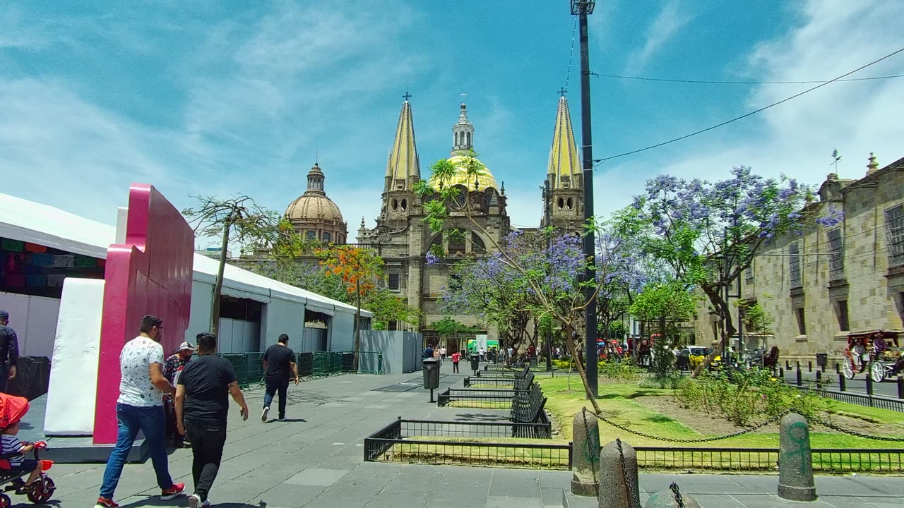 People walk past benches and trees toward a cathedral with golden spires.