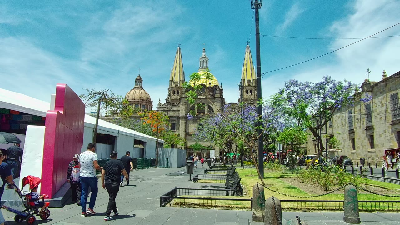 Pedestrians, including an adult pushing a stroller, pass the pink structure in Guadalajara, Mexico, with the cathedral domes and golden spires visible beyond the plaza trees.
