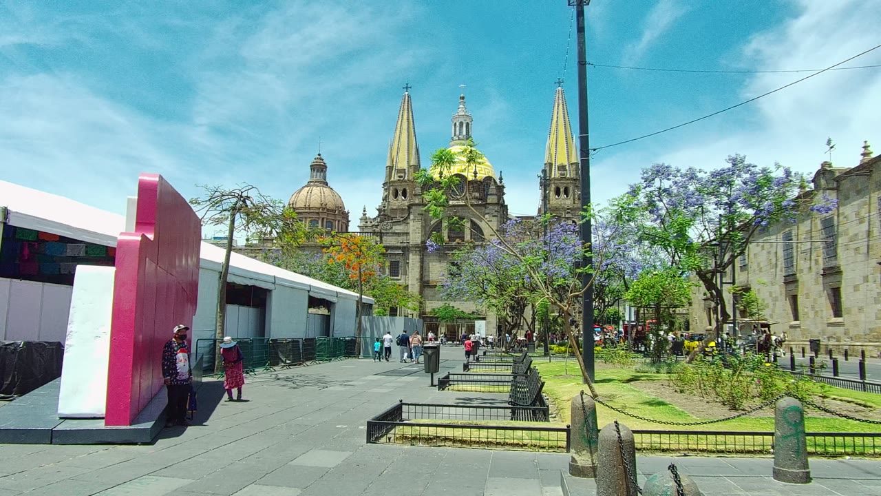 A wide view in Guadalajara, Mexico shows a stone-paved square with a pink and white structure at left, trees and grass in the center, pedestrians, and cathedral domes and spires in the background.