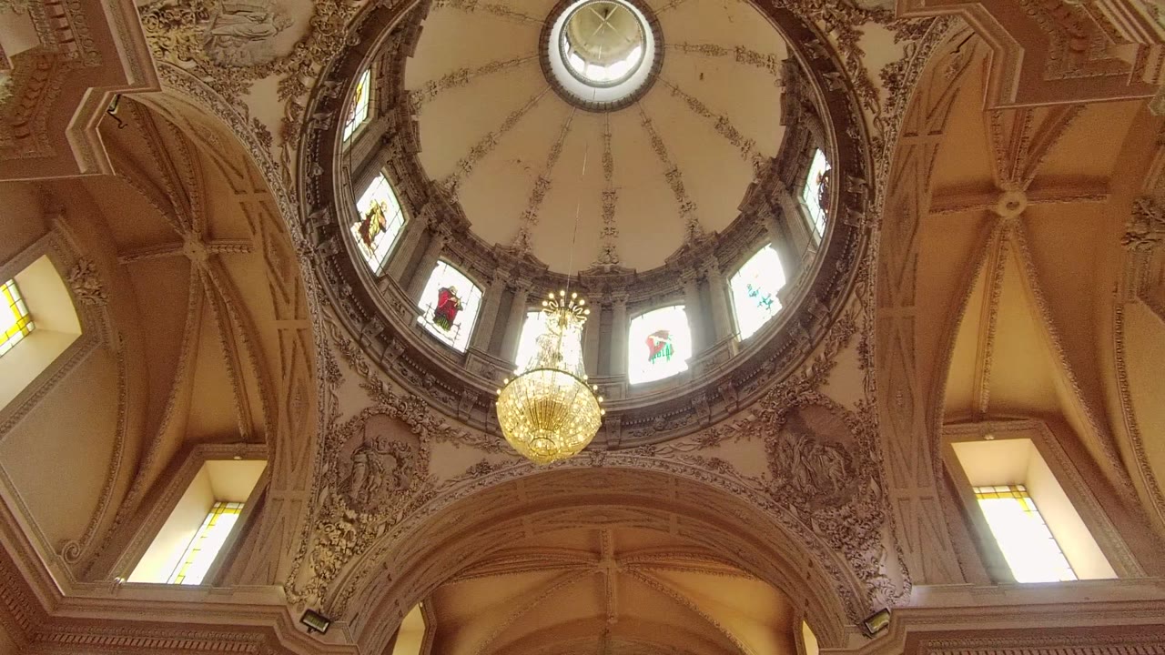 Upward view of an ornate domed ceiling in Guadalajara, Mexico, with a gold chandelier below stained glass windows and no visible people.
