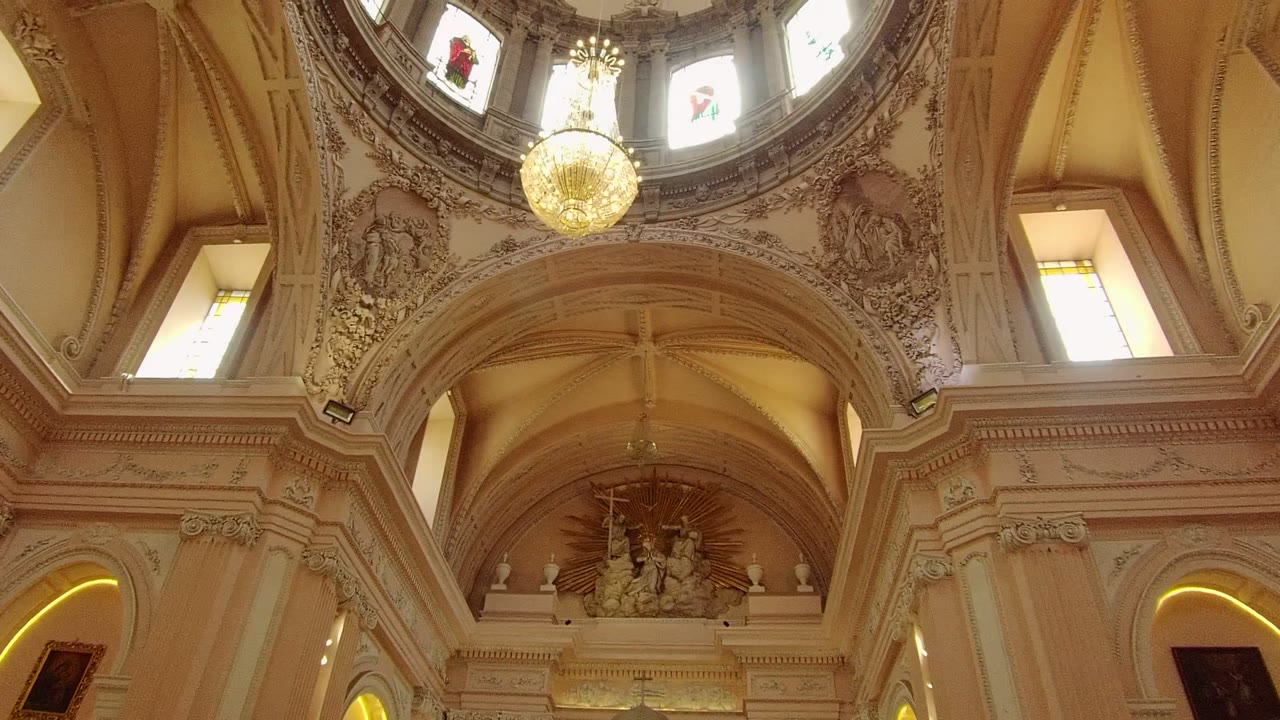 Upward Guadalajara church view with lit arched niches, stained glass panels, carved ceiling decoration, religious wall art, and no visible people.