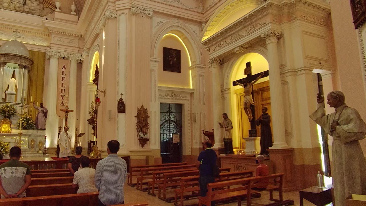 Guadalajara church interior at the start of the clip, with seated worshippers in wooden pews facing a lit altar, statues, candles, and a large crucifix to the right.