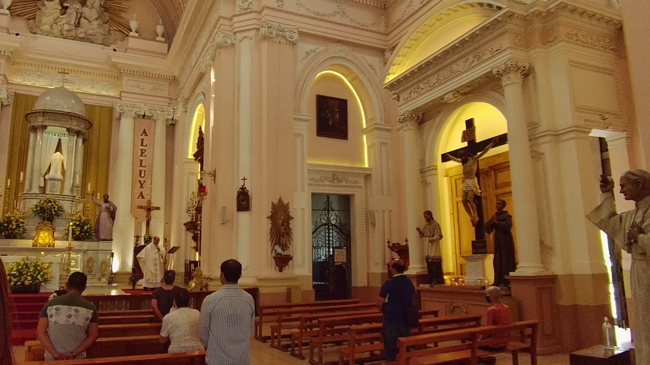 Church nave with people in pews, a lit altar, banners, statues, and a crucifix.