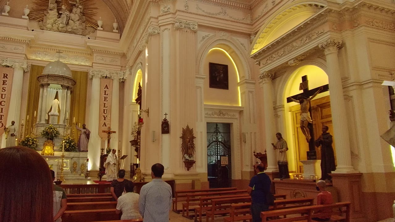Guadalajara church interior at 0:02, with a person's long hair entering the lower foreground while pews, worshippers, banners, statues, and the crucifix remain visible.