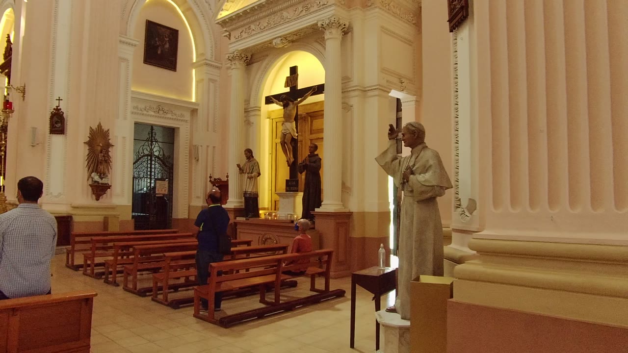 Guadalajara church interior at the start of the clip, with wooden pews, a seated man in the foreground, another visitor near the aisle, white columns, statues, and a large crucifix.