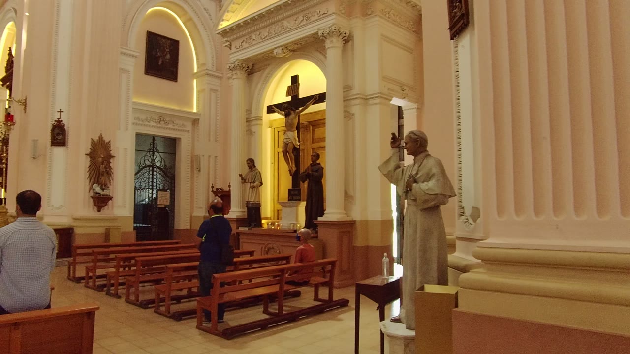 People sit in wooden pews facing religious statues and a crucifix inside a church.