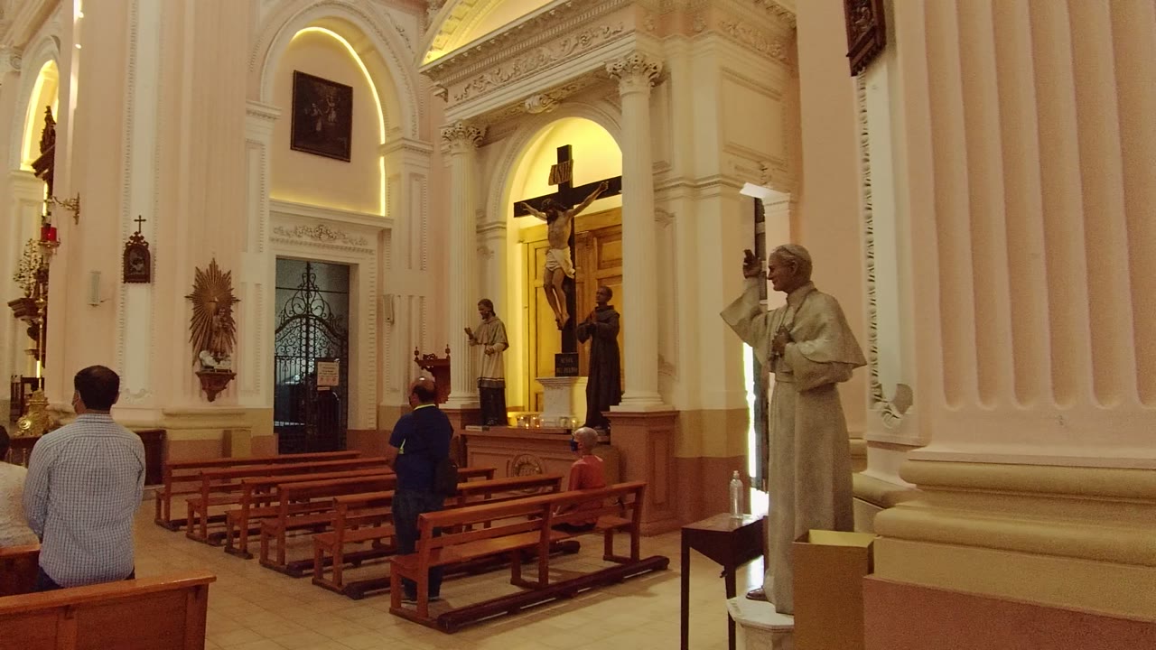 Guadalajara church interior two seconds in, showing the pew rows from a slightly shifted moment with visitors seated below tall white columns and a crucifix lit in a rear niche.