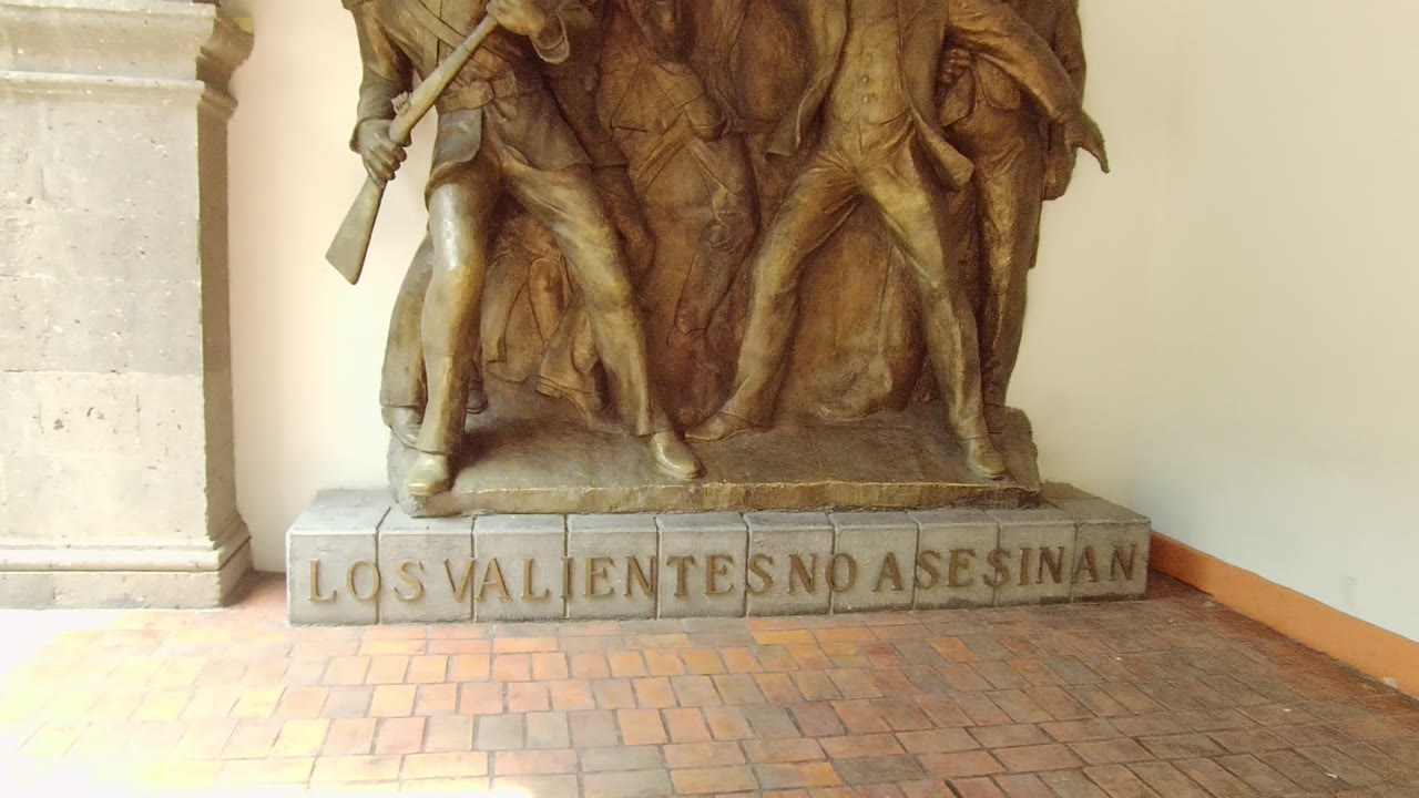 Early frame of a bronze statue in Guadalajara with the stone inscription partly centered, pale columns at left, brick paving below, and no visible people.