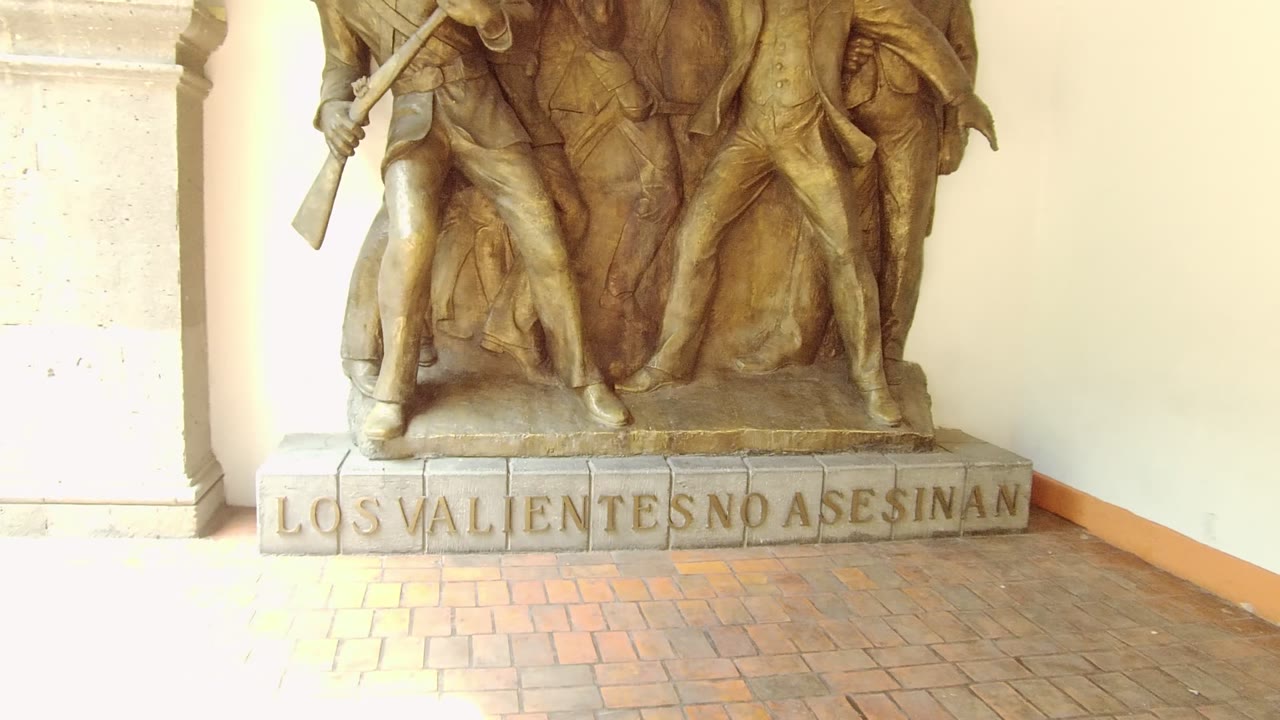 The static shot holds on the Guadalajara memorial statue with warm bronze figures, pale architectural columns, a cream wall, brick paving, and no visible people.